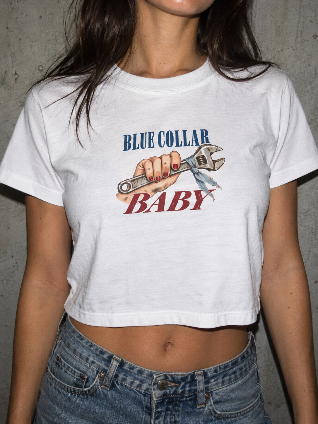 A woman wearing a white cropped graphic t-shirt with 'BLUE COLLAR BABY' and an wrench illustration, paired with blue jeans against a gray concrete wall.