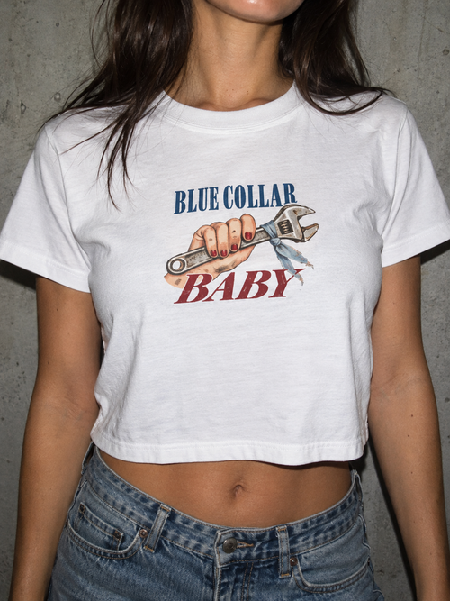 A woman wearing a white cropped graphic t-shirt with 'BLUE COLLAR BABY' and an wrench illustration, paired with blue jeans against a gray concrete wall.