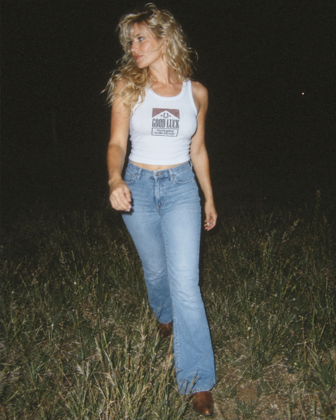 A woman walking outdoors at night in a grassy field, wearing a white tank top with a 'SHOOT BACK' graphic and light blue jeans.