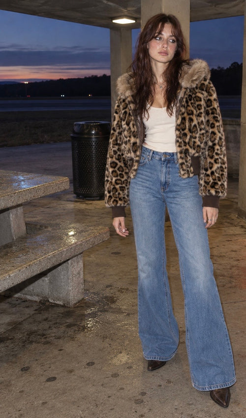 A woman stands under a shelter at dusk wearing a leopard-print faux fur jacket with a white tank top and light-wash flare jeans.