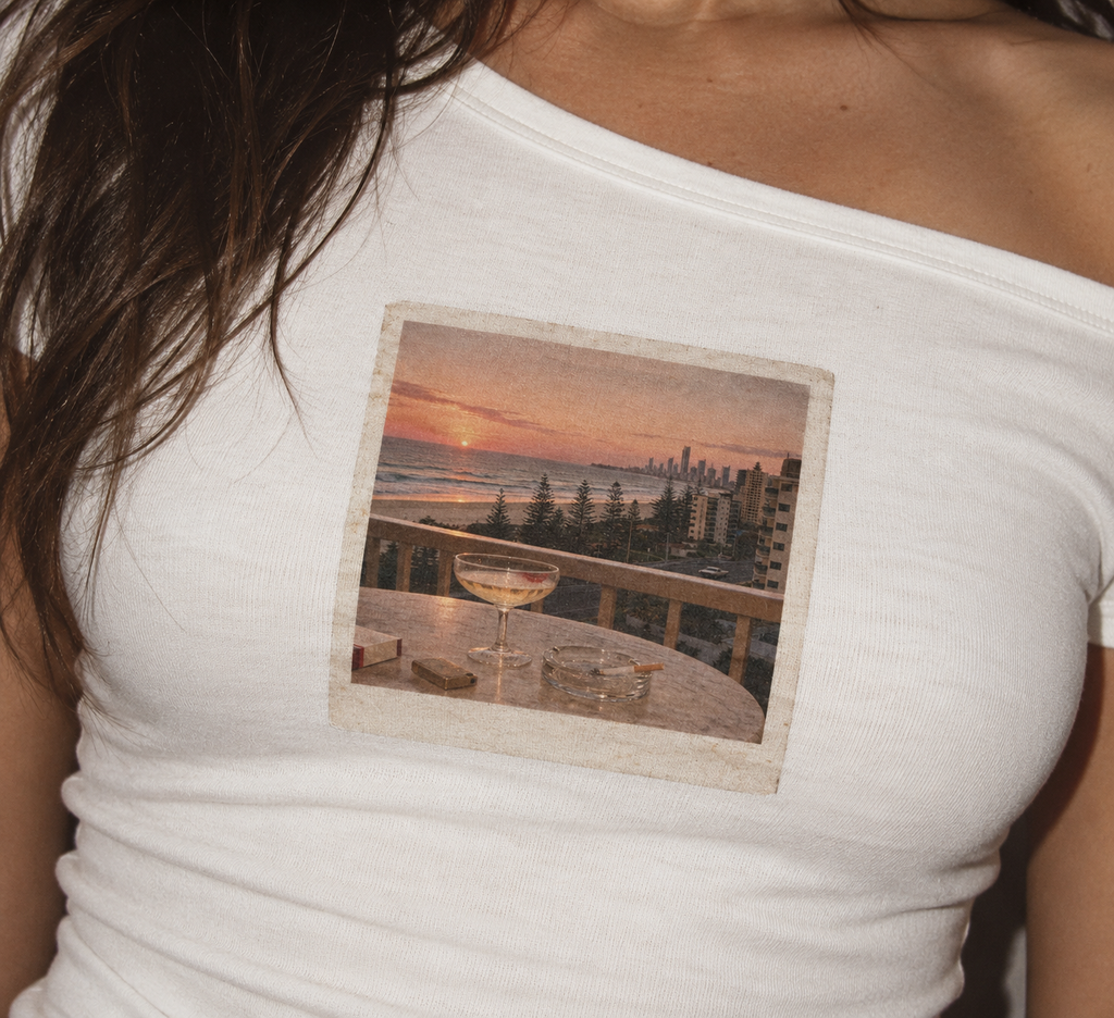 A close-up of a woman wearing a white off-the-shoulder t-shirt with a square graphic print on the chest depicting a sunset beach scene with a wine glass and city skyline.