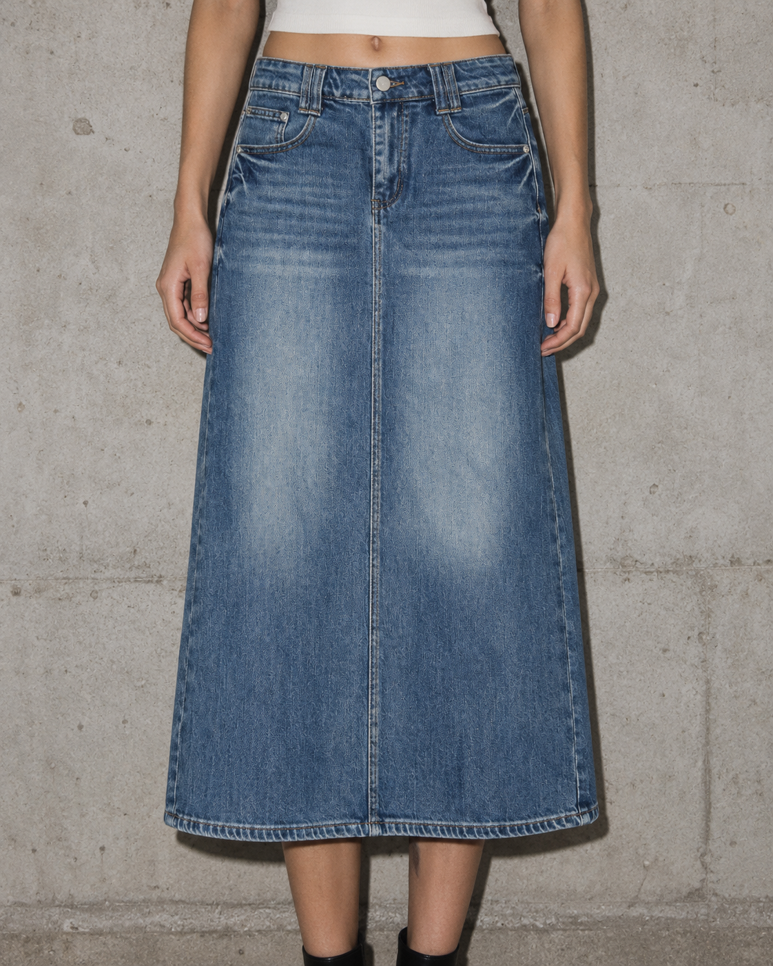 A model wearing a mid-length blue denim skirt with a front fly and five-pocket design, standing against a textured concrete wall.