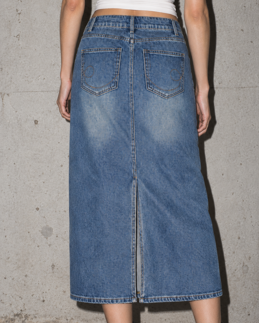 A person wearing a medium-wash blue denim midi skirt with a front slit, shown from the back against a concrete wall.