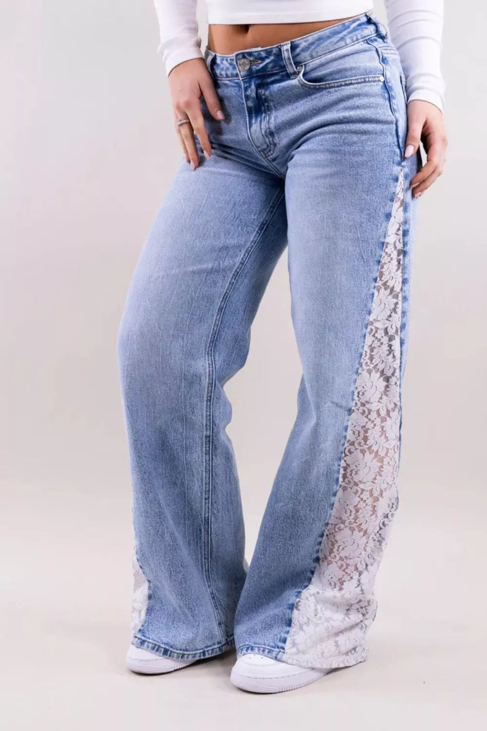 A person wearing light blue wide-leg jeans with white lace panels running down the outer side of each leg, paired with a white long-sleeve crop top and white sneakers, photographed against a plain light studio background.