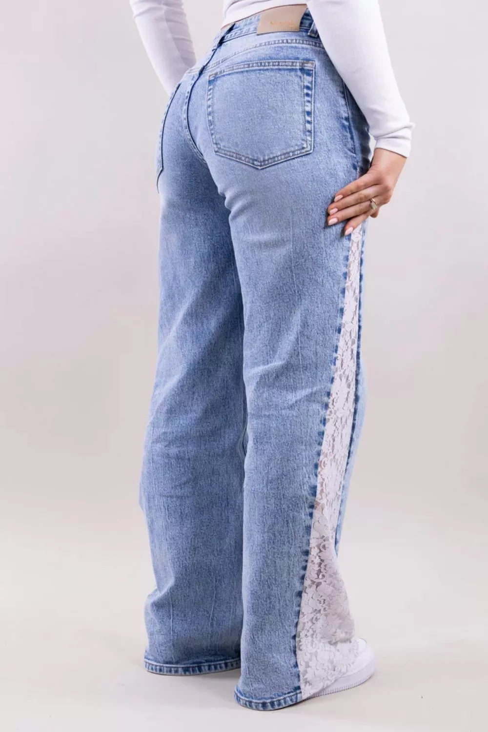 A person wearing light blue wide-leg jeans with a white lace panel down the side, shown from the back against a plain light beige studio background.