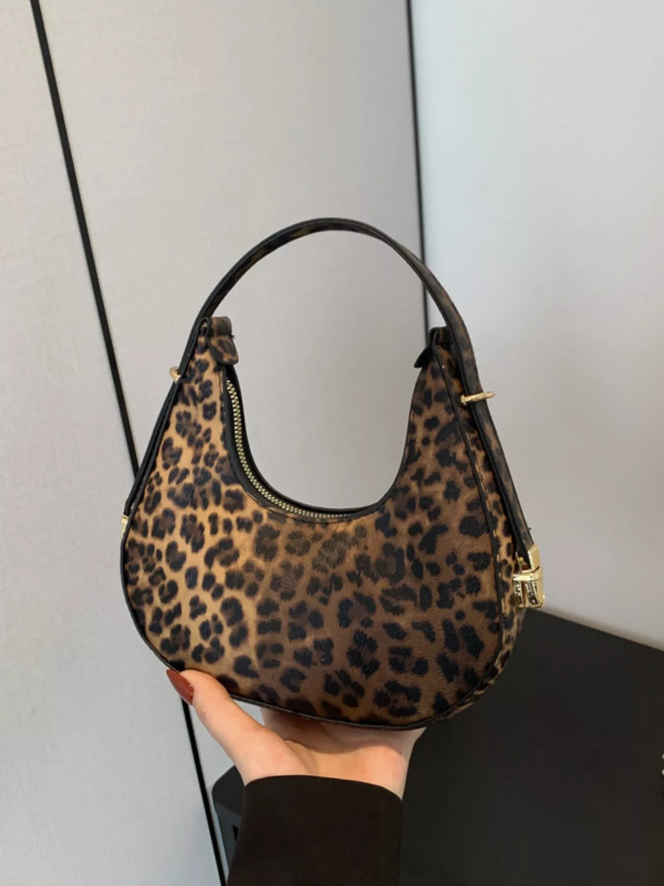 A leopard print shoulder bag with a curved top handle, held by a hand. The bag features a brown and black animal print pattern with gold-tone hardware.