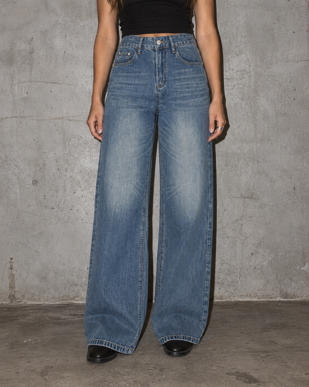 A person wearing high-waisted, wide-leg blue denim jeans with a faded wash, paired with a black crop top and black shoes, standing against a textured gray concrete wall.