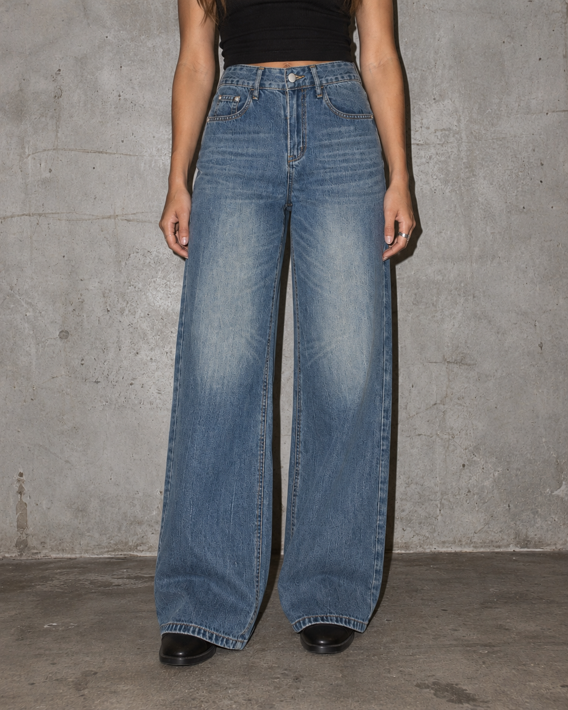 A person wearing high-waisted, wide-leg blue denim jeans with a faded wash, paired with a black crop top and black shoes, standing against a textured gray concrete wall.