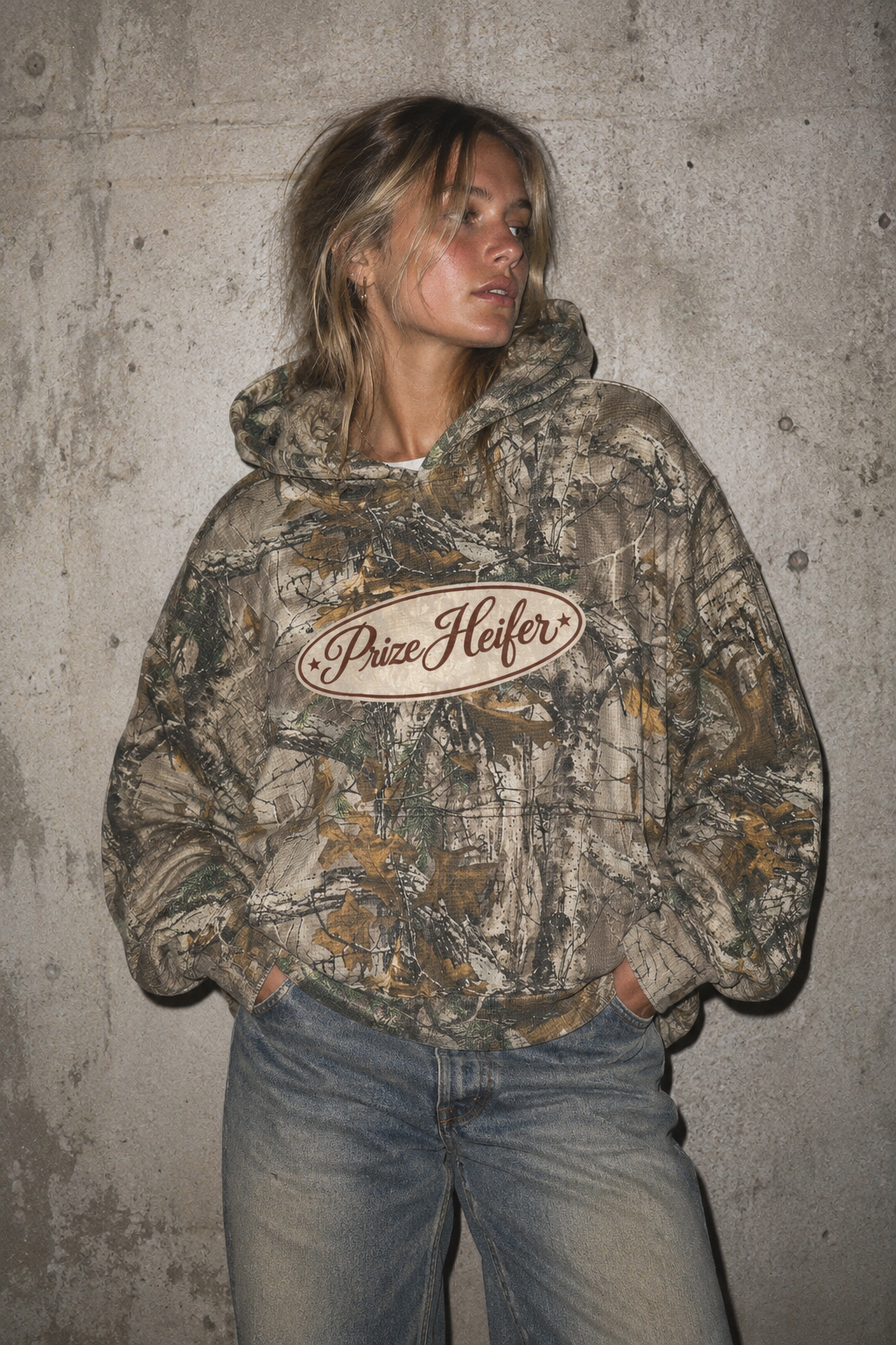 A woman wearing a camouflage hoodie with a 'Price Heifer' logo on the chest, paired with light-wash blue jeans, standing against a textured concrete wall.