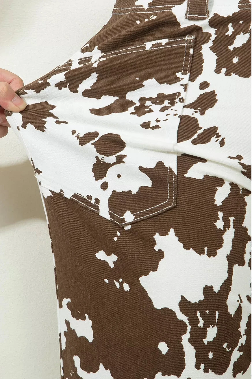 A close-up of a brown and white cow-print garment being held by a hand to show the fabric texture and stretch, featuring a chest pocket with white contrast stitching.