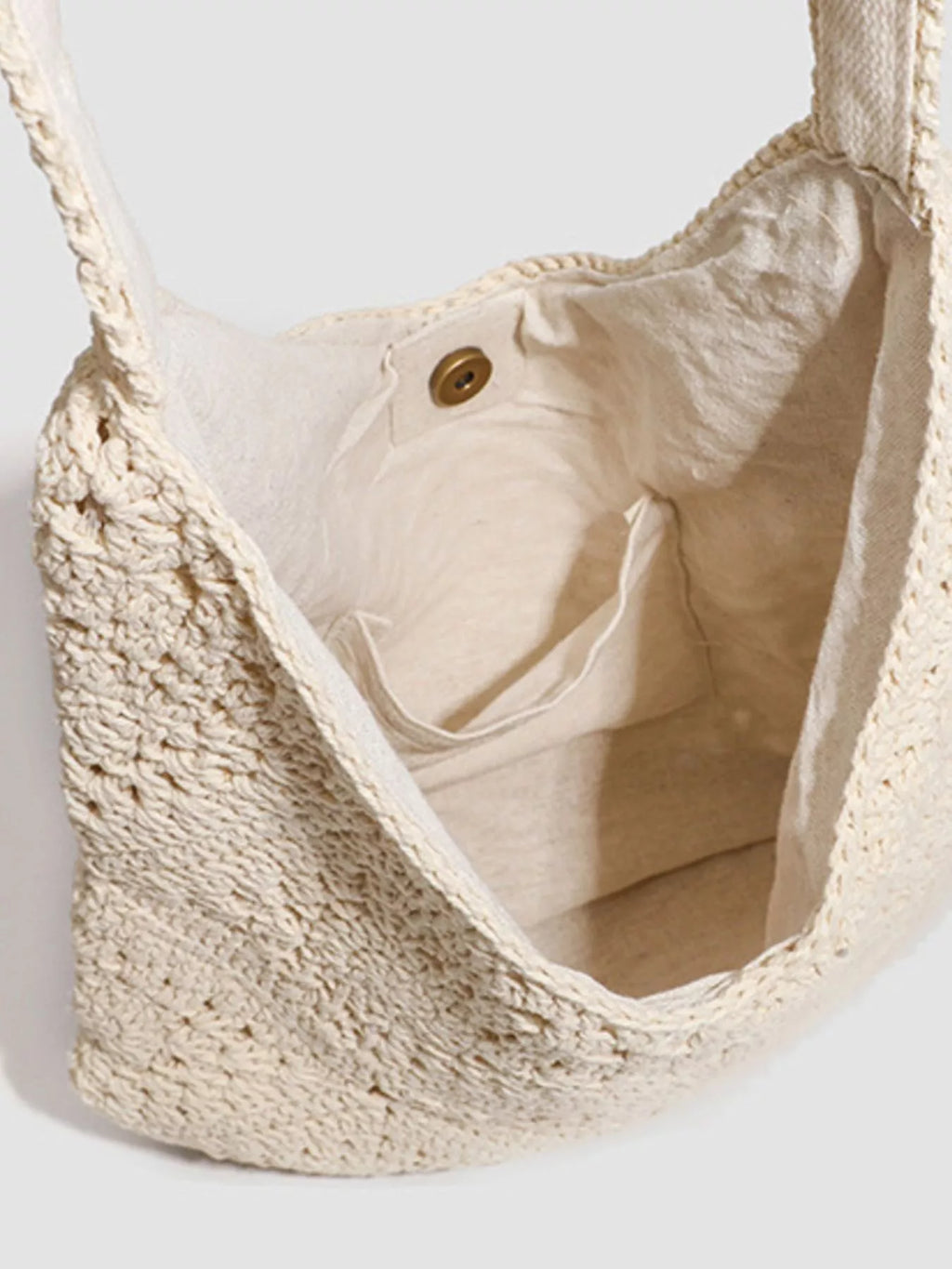 A close-up of a cream-colored crochet shoulder bag with a matching woven strap, showing the textured openwork crochet pattern and the cream fabric lining with a brass snap button.